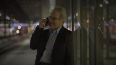 Gülümseyen işadamı, gece bir tren istasyonunda telefon görüşmesi yapıyor. Arka planda şehrin ışıkları var..