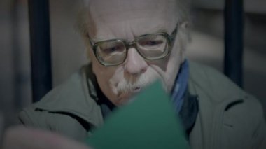 Gözlüklü yaşlı adam belge okurken odaklanmış görünüyor, ciddi bir ifade derin düşüncelere daldığını gösteriyor.
