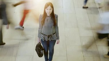 İnsanlar yanından geçerken, modern bir alışveriş merkezinde hareketsiz duran bir kadın kalabalıkların arasındaki yalnızlığı simgeliyor..