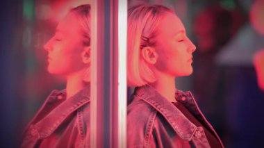 Şık giyinmiş genç bir kadın, kapalı gözlerle, şehir manzarasında pembe neon ışığın altında yansıyor.