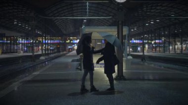 Loş ışıklı tren istasyonunda şemsiyeli iki siluet anonimlik ve yalnızlık dolu sinematik bir atmosfer yaratıyor.