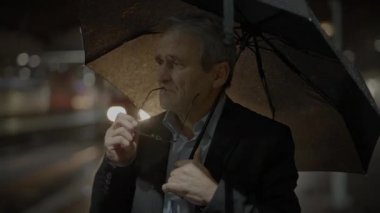 Geceleri şehirde bir adam şemsiyenin altında elinde gözlükleriyle dikilir. Arka planda bulanık ışıklar vardır..