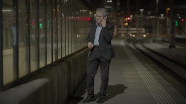 Takım elbiseli bir işadamı gece modern bir tren istasyonunda beklerken telefonla konuşuyor..