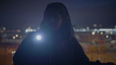 Kapüşonlu siluetli bir şahıs gece boyunca şehir ışıklarının önünde duruyor ve gizemli bir sahne yaratıyor..