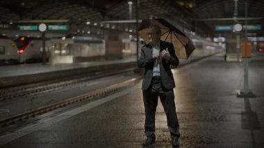 Elinde şemsiye tutan bir işadamı, arka planda trenlerle yağmurlu bir tren istasyonunda bekler..