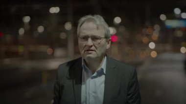 Gece şehir merkezinde takım elbiseli yaşlı bir adam tek başına duruyor. Arka planda bulanık şehir ışıkları var.