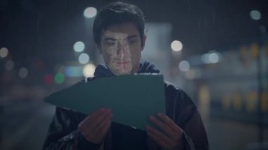 Genç adam, yağmurda sokak lambalarının altında mektup okuyor. Düşüncelere dalmış, gece vakti şehir ortamında..