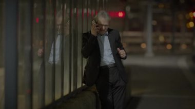 Takım elbiseli bir işadamı gece dışarıda, arka planda şehir ışıkları olan cam bir duvarın yanında telefonla konuşuyor..