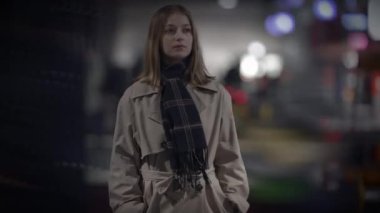 Trençkot ve eşarp giyen zarif bir kadın bulanık bir arka planı olan bir şehir gece sahnesinde duruyor.
