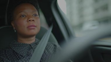 Arka planda bulanık binalar olan bir şehirde derin düşüncelere dalmış bir kadının araba sürüşü..
