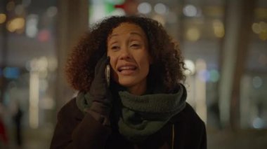 Bir kadın gece şehirde bir telefon görüşmesi yapıyor, arkasında bulanık şehir ışıkları olan bir ceket ve eşarp giyiyor.