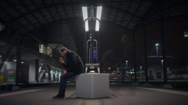 Geceleyin modern bir tren istasyonunda tek başına oturan, ışıklarla aydınlanan, yalnızlık çeken bir adam..