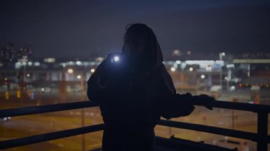 Geceleri çatıda feneri olan gizemli bir figür, arka planda şehir ışıkları, gizemli bir ortam yaratıyor.