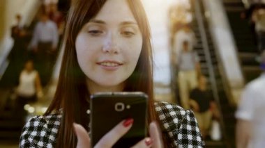 Telaşlı bir alışveriş merkezinde akıllı telefon kullanan gülümseyen genç bir kadın. Yürüyen merdivenler ve arka planda yürüyen insanlar.