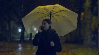 Yağmurlu bir gecede loş bir parkta bir kadın cep telefonuyla konuşurken ve şemsiyesini tutarken yürür.