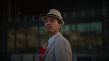 Bej şapka ve ceket giyen şık bir adam arka planda bulanık bir şekilde şık bir şekilde poz veriyor.