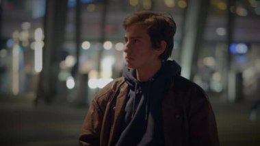 Güz kıyafetleri içinde genç bir adam arka planda bulanık şehir ışıklarıyla hareketli bir şehirde duruyor..