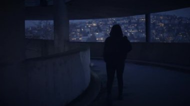 Kapüşonlu ceketli bir şahıs, loş ışıklı çok katlı bir binada gece yürürken gizemli bir atmosfer yaratıyor.