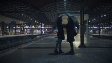 İki siluetli şahıs şemsiye tutuyor ve yağmurlu bir gecede tren istasyonunda sohbet ediyorlar..