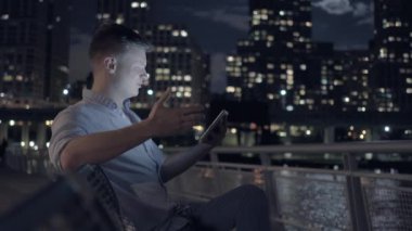 Geceleri dijital tabletle meşgul olan bir adam, şehir bankında oturuyor, parlayan bir silueti var..