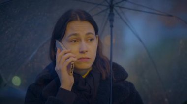 Yağmurlu bir şehirde genç bir kadın şemsiyeyle cep telefonuyla konuşuyor, arka planda bulanık bokeh ışıkları var.