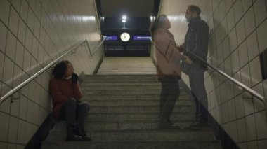 Kıvırcık saçlı bir kadın gece metro merdivenlerinde oturur ve arka planda bir saat görünür.