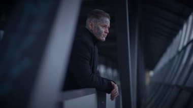 Koyu renk paltolu adam korkuluklara yaslanıyor, modern bir mimari koridorda, düşük ışıklandırmalı....