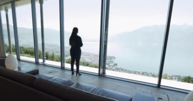 Bir kadın, modern bir evde büyük pencereler önünde dururken sakin bir göl ve dağ manzarasının tadını çıkarıyor.