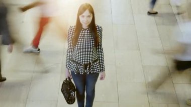 Genç bir kadın hareketsiz duruyor, modern bir koridorda bulanık hareketlerle çevrili, tefekküre sembolik..