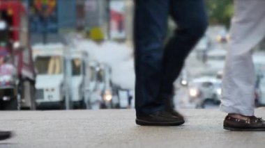 Arka planda trafik ve şehir hayatı olan kalabalık bir şehir caddesinde yürüyen yayaları gösteren bir sokak sahnesi.