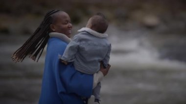 Bir anne çocuğunu mavi bir battaniyeye sarmış, açık bir derenin yanında hassas bir anı paylaşıyor..