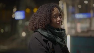 Kıvırcık saçlı ve eşarplı bir kadın, geceleri açık havada derin derin düşünür, şehirde huzurlu bir atmosfer yaratır.