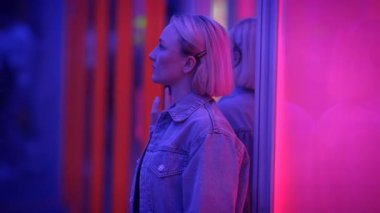 Kot ceketli kısa sarı saçlı şık bir kadın neonlit bir odada cama yaslanıyor.
