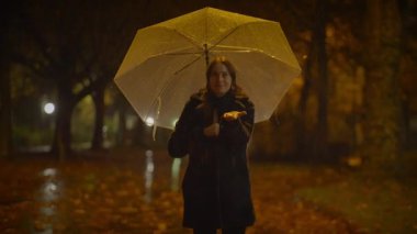 Bir kadın elinde şemsiyeyle yağmurlu bir parkta dikilip karamsar, atmosferik bir sahne yaratıyor..