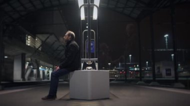 Modern, fütüristik bir tren istasyonunda, bir adam gece yalnız başına bekler, yalnızlık yolculuğunu yansıtır.