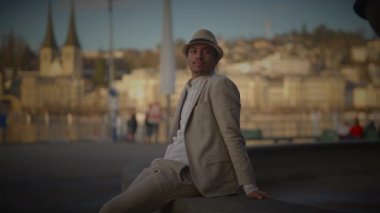 Bej takım elbiseli ve fötr şapkalı bir adam nehir kenarındaki bir çıkıntıya yaslanıyor ve arkasında tarihi binalar var..