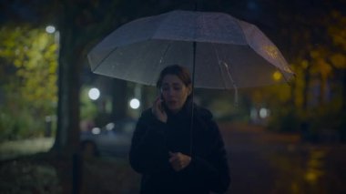 Bir kadın loş ışıklı, yağmurlu bir parkta şemsiyenin altında telefonla konuşarak sakin bir atmosfer yaratıyor.