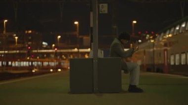 Geceleyin loş bir tren istasyonundaki bankta oturan bir adam huzurlu ve düşünceli bir atmosfer yaratıyor.