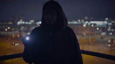 Kapüşonlu ve el fenerli gizemli bir figür gece bir şehir manzarasına karşı duruyor, belirsizlik yaratıyor.