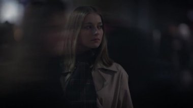 Uzun saçlı genç bir kadın gece bir şehir sahnesinde duruyor, ceket ve eşarp giyiyor, karamsar bir atmosfer yaratıyor.