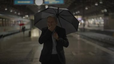 Yağmurlu bir gecede, loş ışıklı bir tren istasyonunda, şemsiyeli yalnız bir şahıs yüzünü gizliyor.