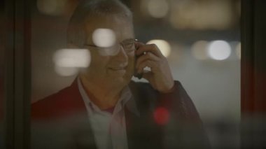 Yetişkin bir iş adamı gece telefonda konuşuyor. Şehir ışıklarının yansımalarıyla camdan görülüyor.