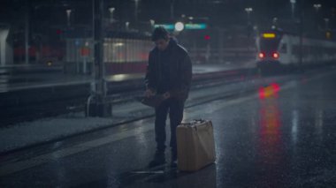 Bavullu yalnız bir adam yağmurlu istasyon platformunda durur, telefonuna bakar, kendini yalnız hisseder.