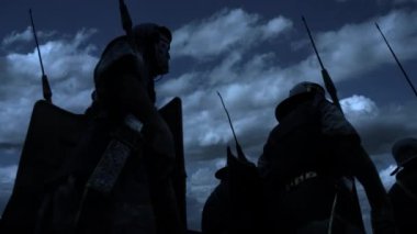 Siluetli ortaçağ savaşçıları bulutlu bir gökyüzü arka planına karşı ellerinde mızrak ve kalkanlarla tasvir edilir.