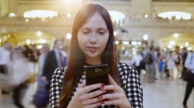Bir kadın kalabalık bir tren istasyonunda akıllı telefonunu kullanarak modern şehir yaşamını somutlaştırıyor.