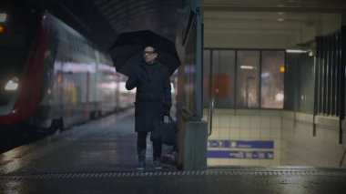 Yağmurlu tren istasyonunda şemsiyeli ve valizli bir adam bekliyor. Soğuk ve nemli bir gecede şehir ışıklarıyla çevrili.