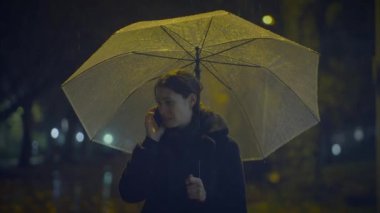 Yağmurlu bir şehir gecesinde şemsiyenin altında telefonla konuşan bir kadın dengesiz bir şehir atmosferi yaratıyor.