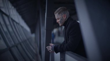 Koyu renk elbiseli bir adam korkuluklara yaslanıyor, modern bir mimari ortamda düşünceli görünüyor..
