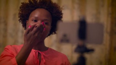 Afro-Amerikalı bir kadın evde bir video bloğu çekiyor, cilt bakımı yapıyor ve akıllı telefon kamerasıyla güzellik içeriği yaratıyor.