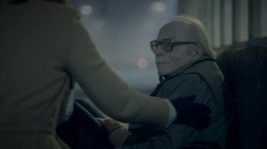 Geceleyin kaldırımda oturan yaşlı bir adam, arka planda kimliği belirsiz bir şahıs yaklaşırken,.